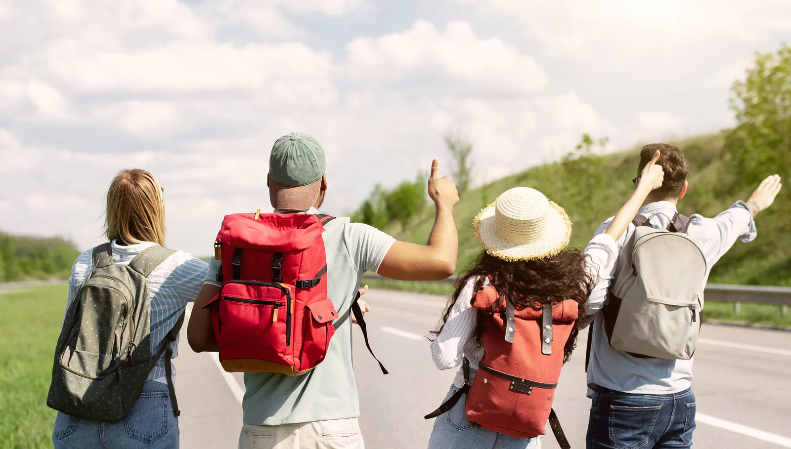 Four backpackers standing beside a rural road with their thumbs raised